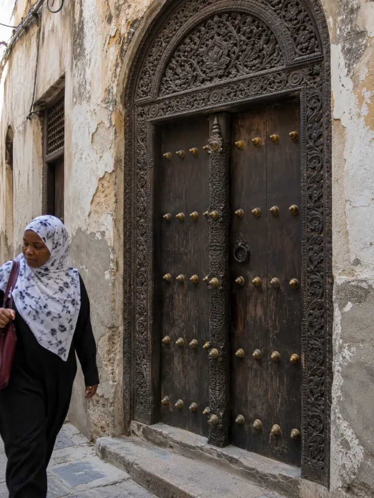 A woman walking past a carved wooden door in a Stone Town alley, Zanzibar